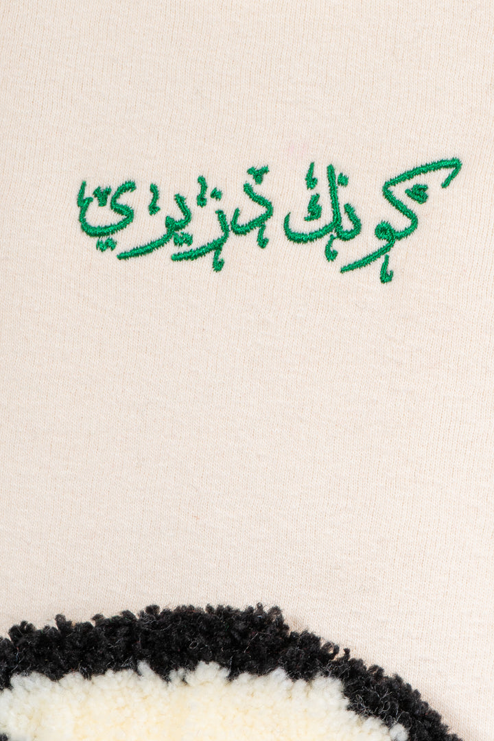 Détail de la broderie verte « Sois Algérien » en arabe sur le pull Numidie beige.
