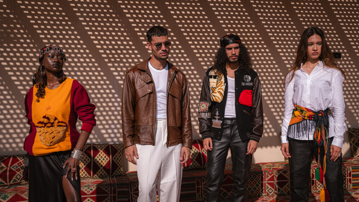 Quatre mannequins portant les créations Numidie : pull Kouffa jaune et bordeaux, veste Azir en cuir marron, Varsity Jacket Tamurt et pantalon en cuir noir, dans un décor artisanal algérien.
