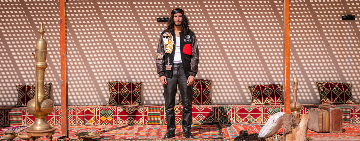 Mannequin portant la Varsity Jacket Tamurt et un pantalon en cuir noir Numidie, dans un décor traditionnel algérien aux tapis berbères.